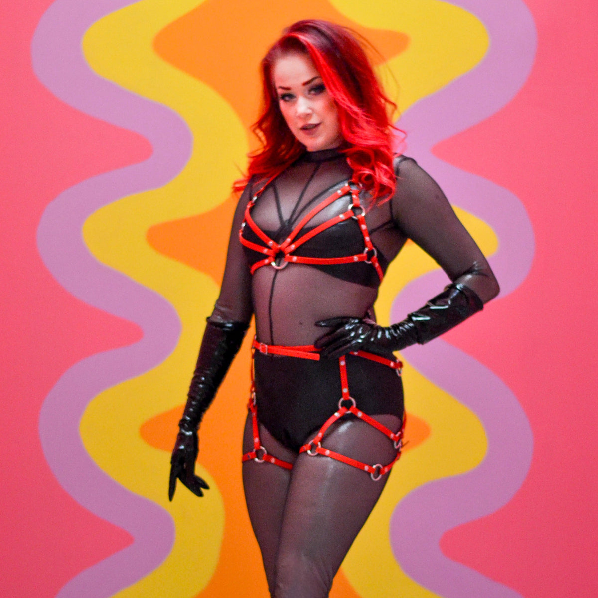 Woman with red hair wearing a black catsuit and a red thigh harness over it.