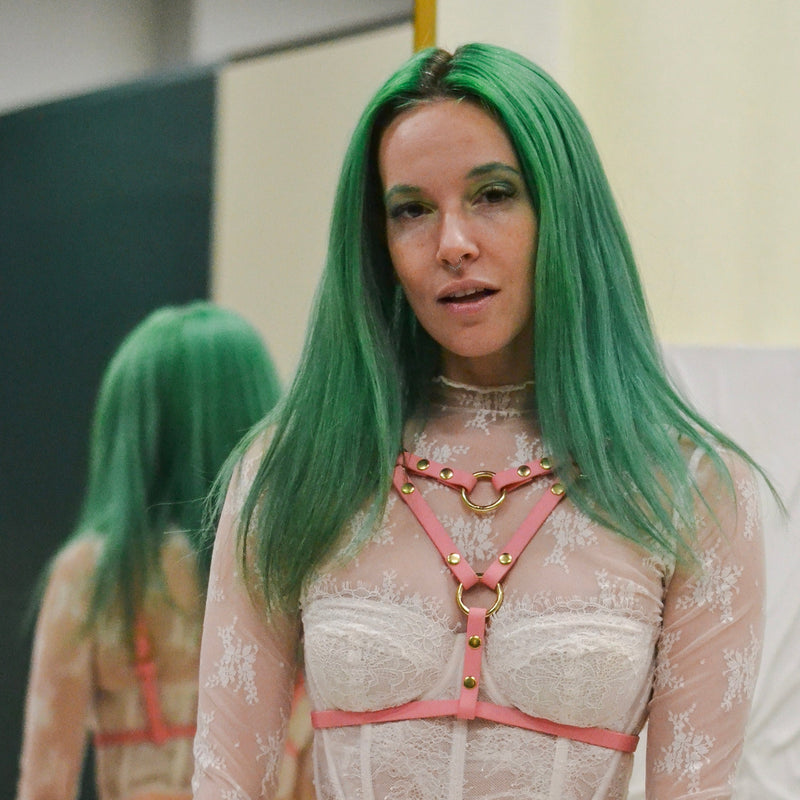 Image of a woman with green hair in a ivory lace bodysuit wearing a angle grinder chest harness in pastel pink.