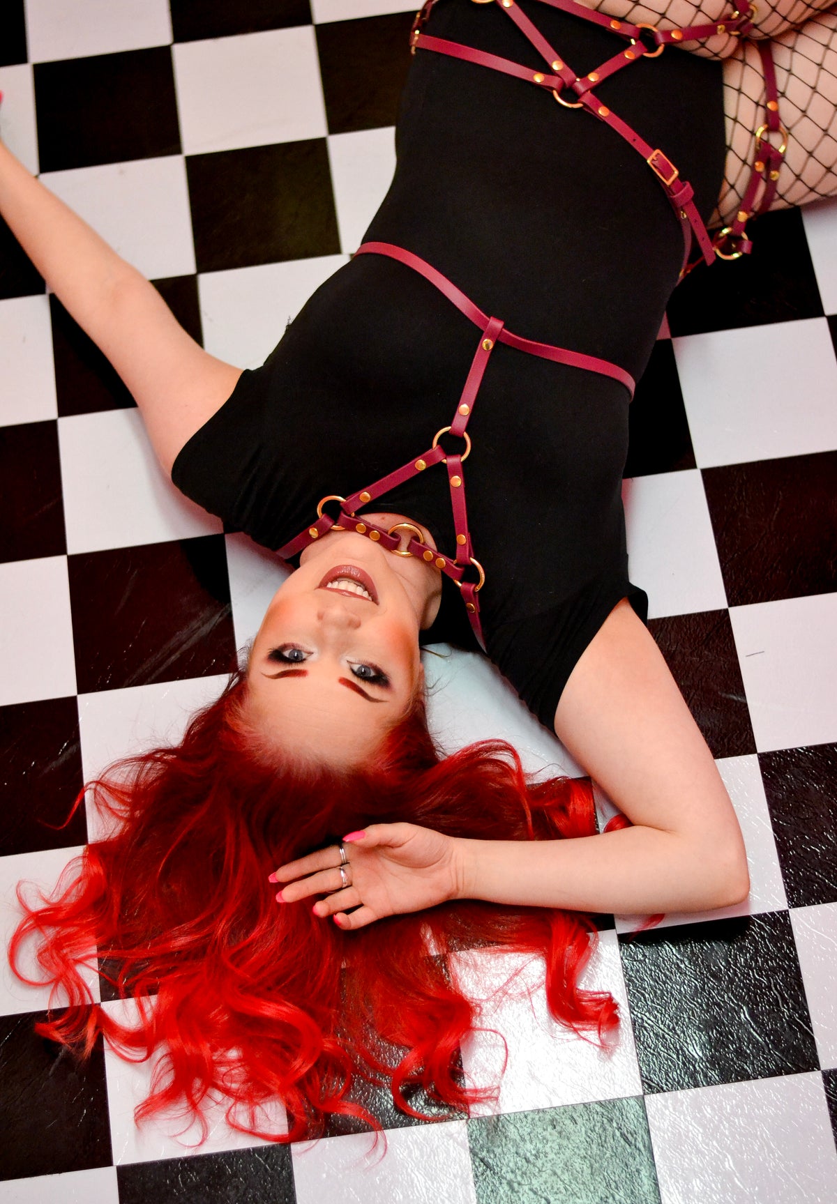 Woman with red hair laying on a checkered floor while wearing a red waist harness over a black bodysuit.