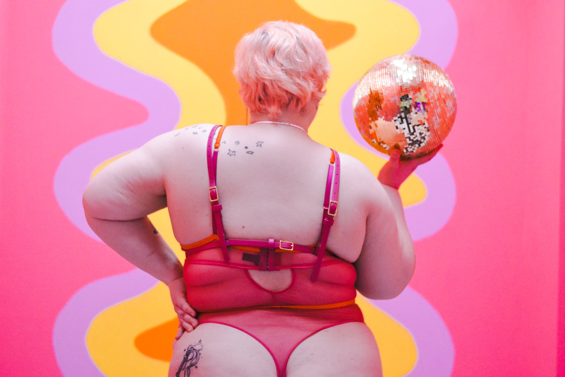 View of person from the back wearing a pink bodysuit and a red harness over it.
