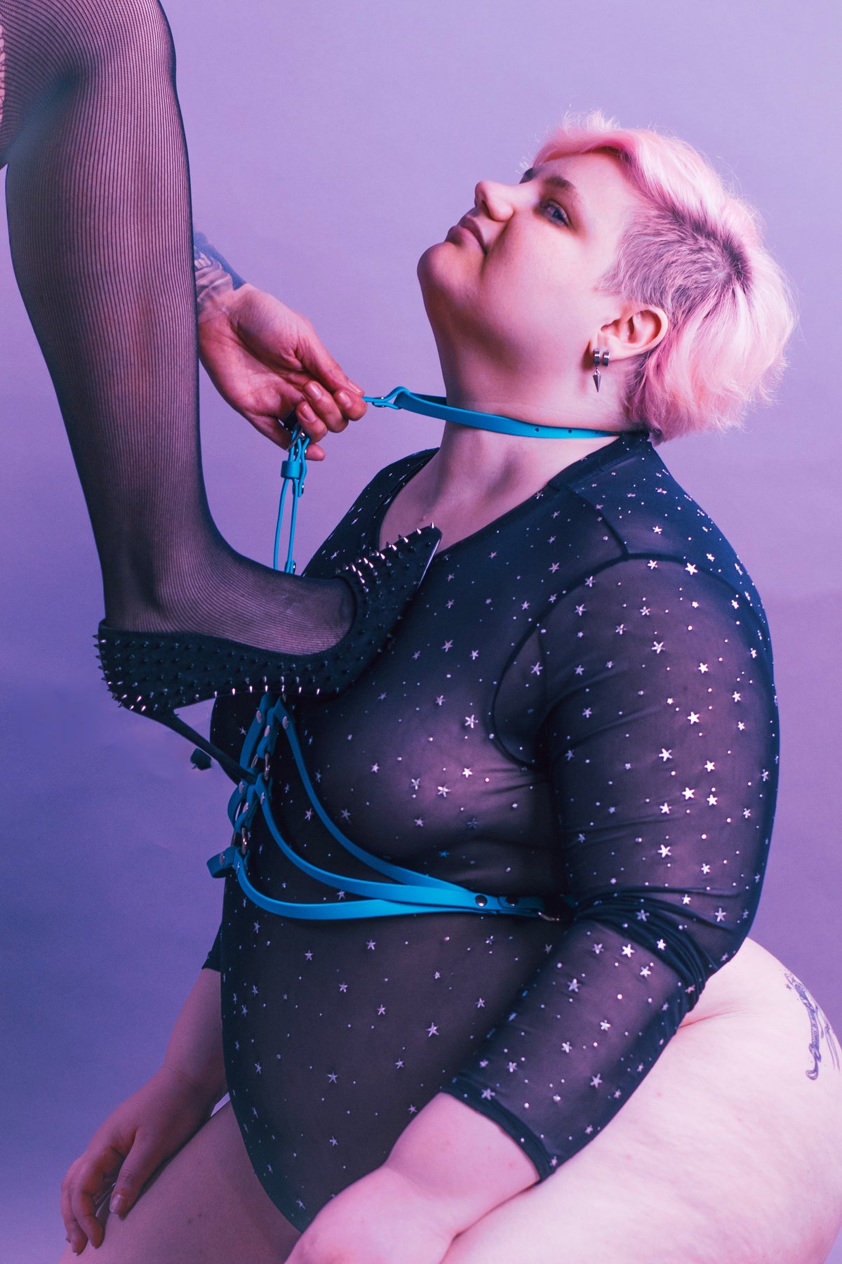 Woman kneeling while wearing a turquoise waist cinch harness as somebody pulls her collar.