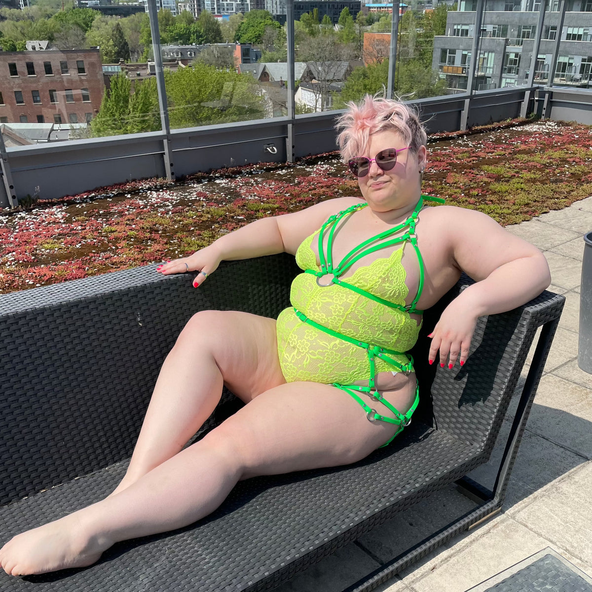 Plus sized woman outside sitting on a couch in a neon bodysuit, a harness top outfit and a sexy butt harness.