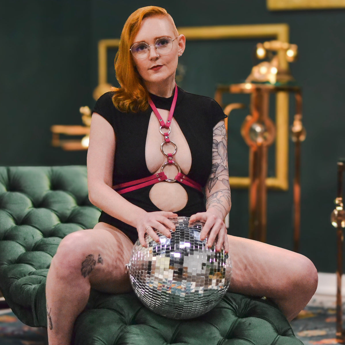 Woman sitting on a green lounger straddling a disco ball showing off a magenta chest harness over her black bodysuit.