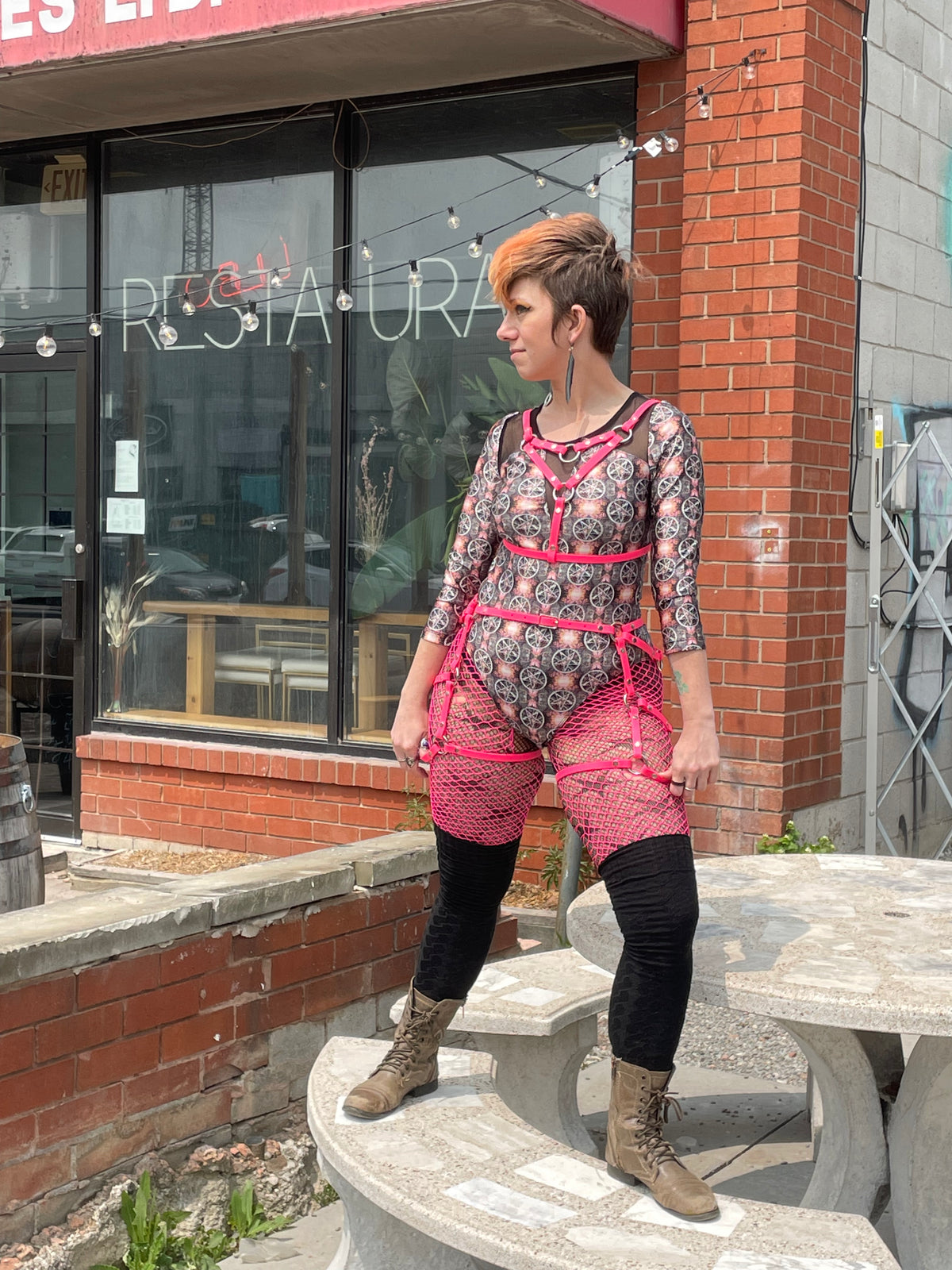 Woman with short hair poses on top of a table wearing a patterned cat suit a pink chest harness bra and thigh harness outfit