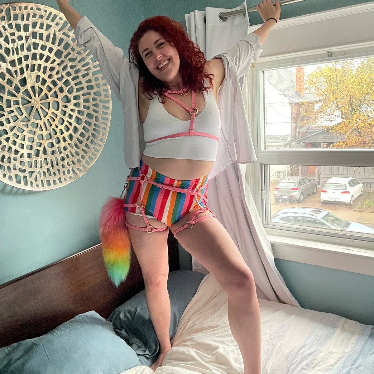 Woman poses on a bed wearing rainbow shorts white crop top pink chest bra harness and thigh harness outfit