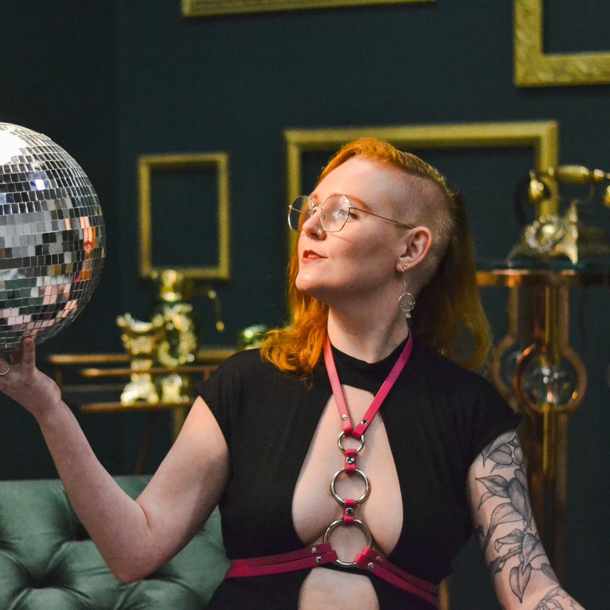 Woman with red hair wearing a magenta circles waist harness while holding a disco ball.
