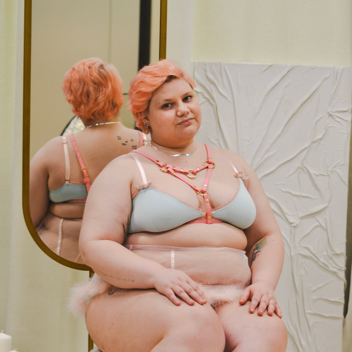 Person with cropped pastel pink hair posing wearing a pastel blue bra top and a pastel pink chest harness.