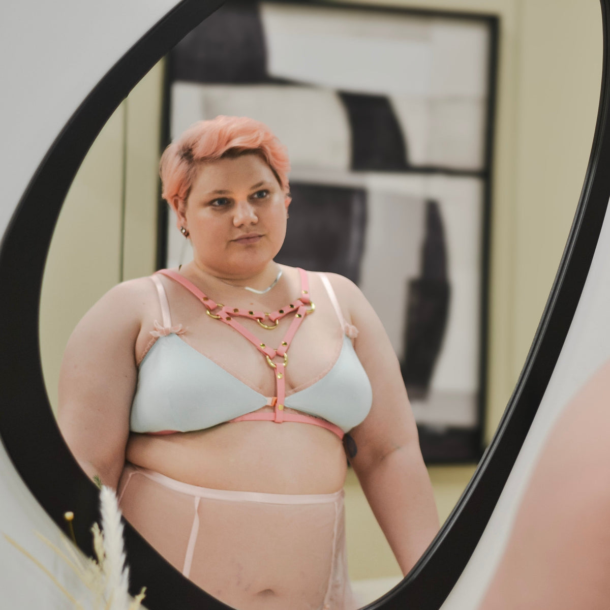 Person with cropped pastel pink hair posing wearing a pastel blue bra top and a pastel pink chest harness while looking at themselves in the mirror.