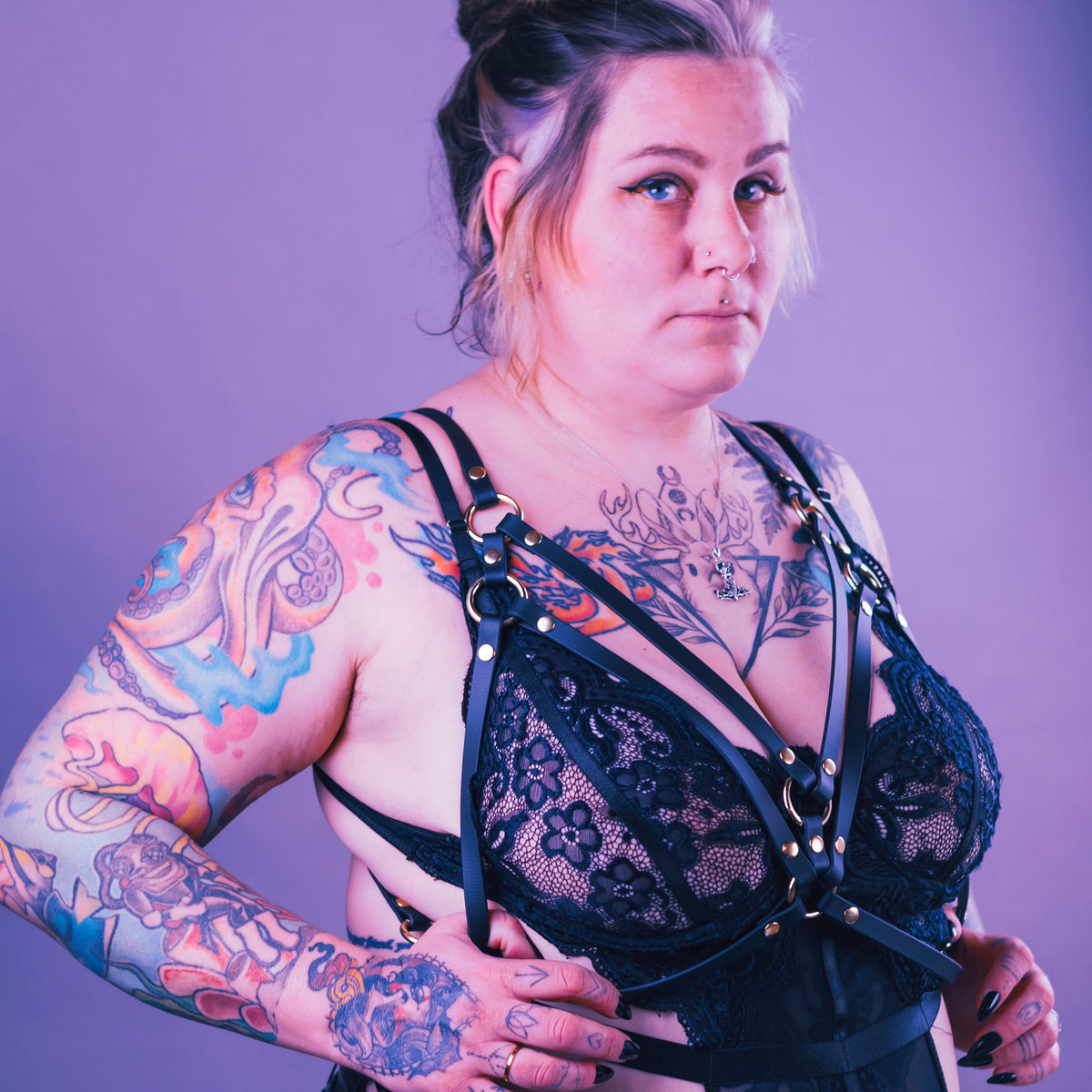 Portrait style image of a heavily tattooed woman wearing black lingerie with a black vegan harness over it.