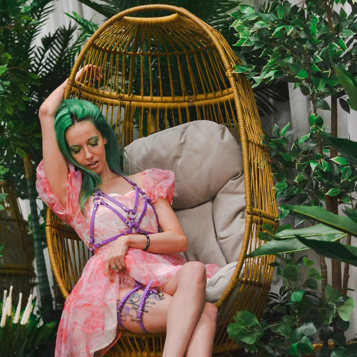 Woman styled on a classic egg chair with a purple reversible lust chest harness and pink dress.