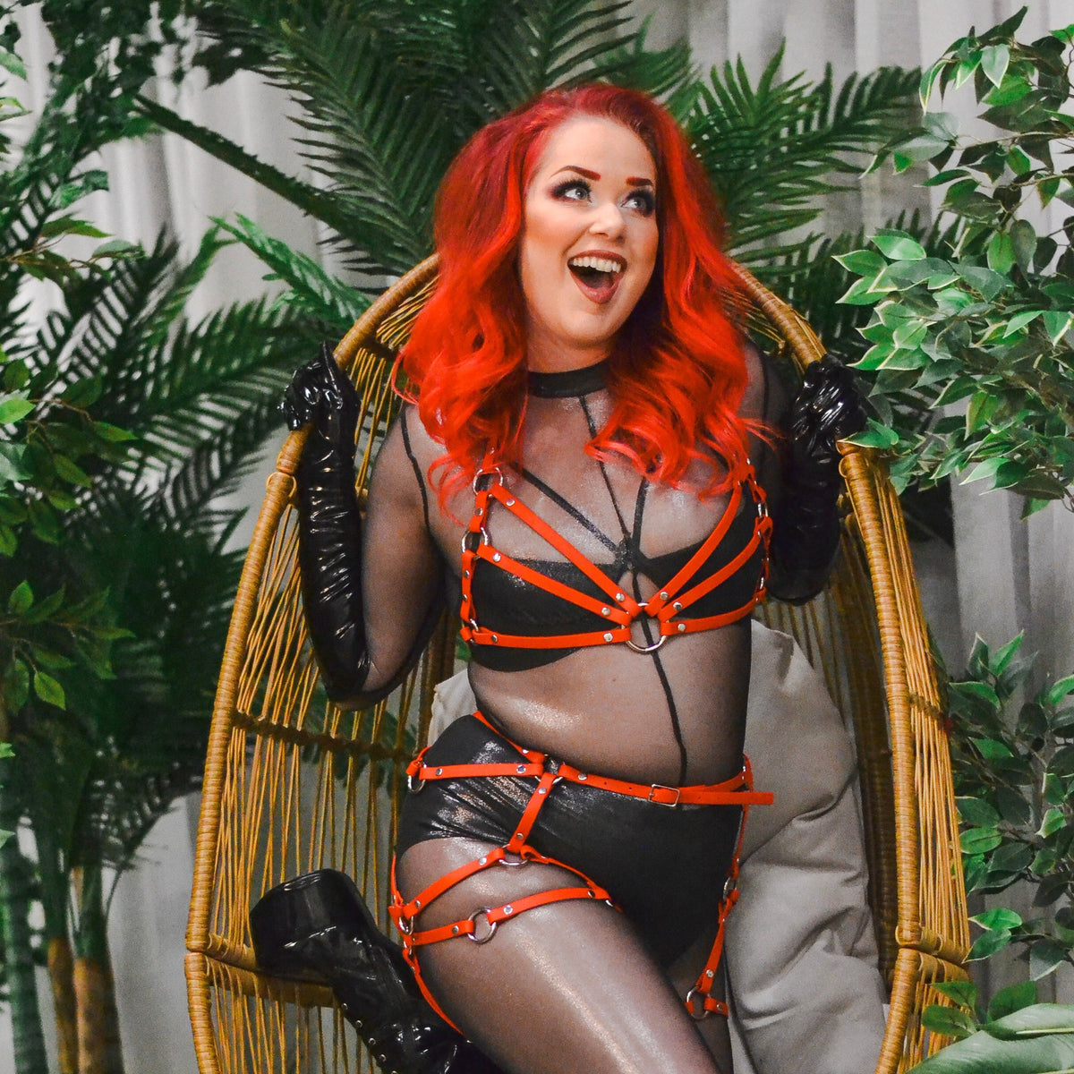 Woman with red hair wearing a black catsuit and a red thigh harness over it.