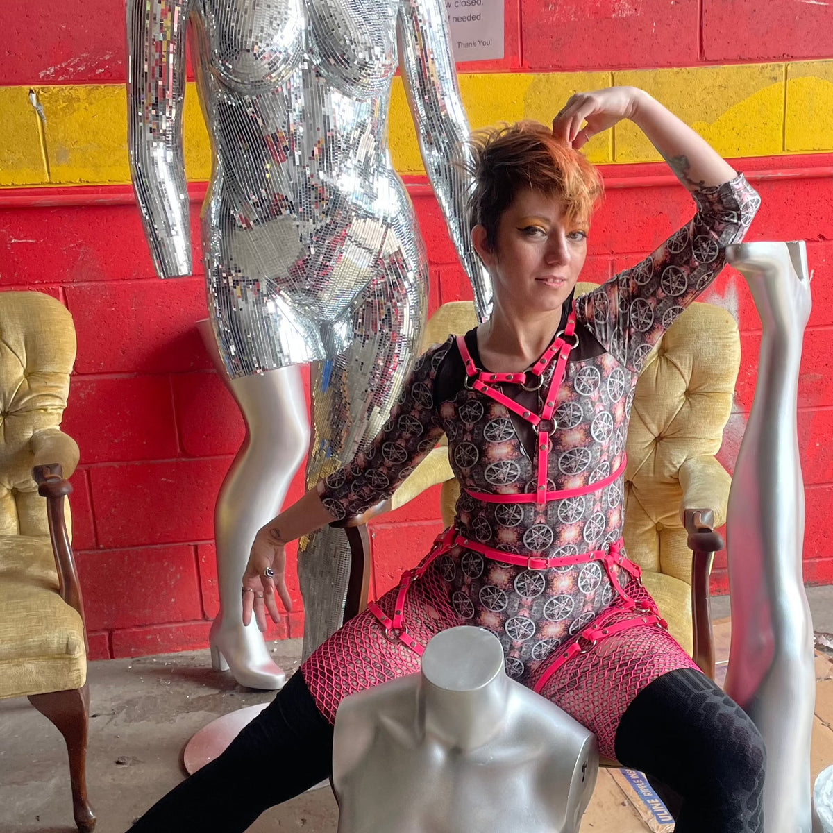 Woman in printed catsuit poses with mannequins in pink chest harness bra and thigh harness outfit.