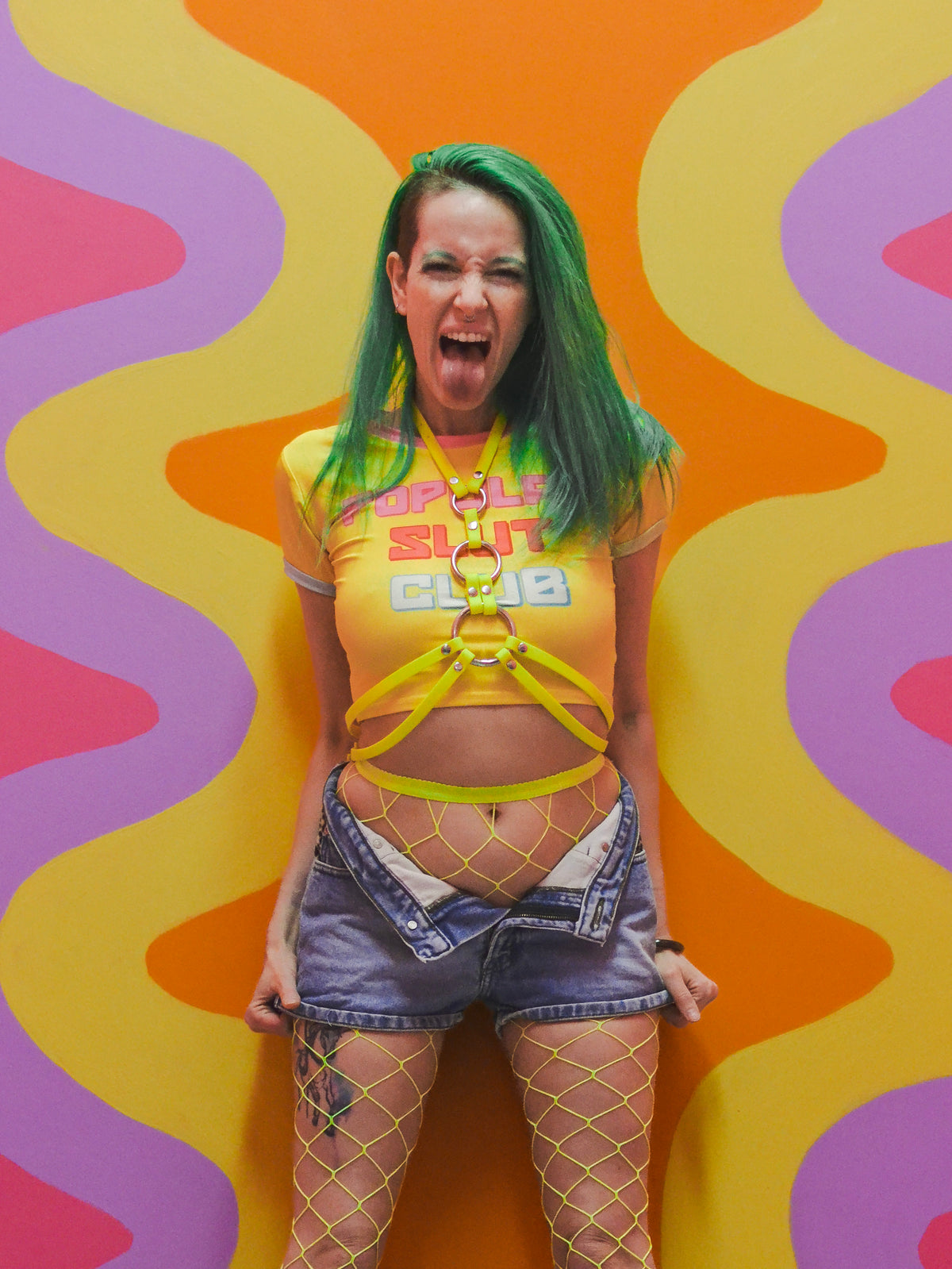 Image of a woman with green hair sticking out her tongue. She is wearing yellow fishnet stockings, short denim shorts, a yellow tshirt, and a neon yellow chest harness.